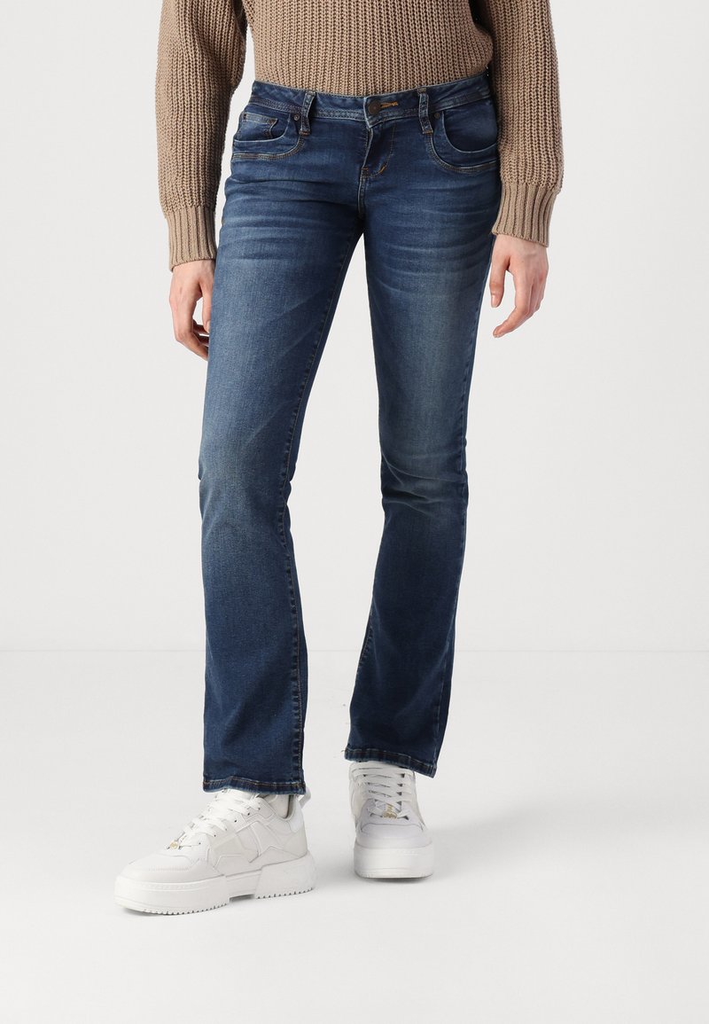 Person in blauen geraden Jeans, weißen auffälligen Sneakers und einem beigen Strickpullover, der auf einem weißen Boden vor einem schlichten Hintergrund steht.