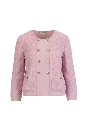 Frau trägt eine strukturierte pinke Jacke mit goldenen Knöpfen, passende pinke Hosen, goldene Schuhe und hält eine beige gewebte Clutch.