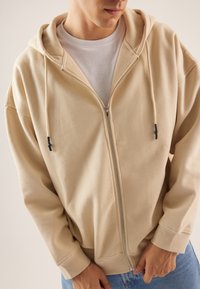 Man in een beige zip-up hoodie over een wit shirt en blauwe jeans, met handen bij de rits in een neutrale achtergrond.