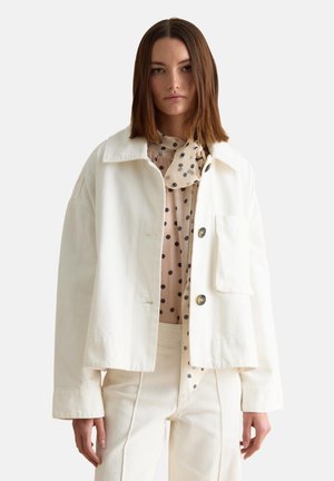 Woman wearing a white oversized jacket over a beige blouse with black polka dots and matching white high-waisted pants, standing against plain background.