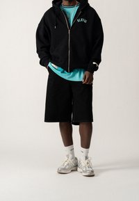 Sorte zip-up hættejakke med "BLK" bogstaver, aqua mesh tanktop, sorte shorts, hvide crew-sokker og grå løbesko med accenter.