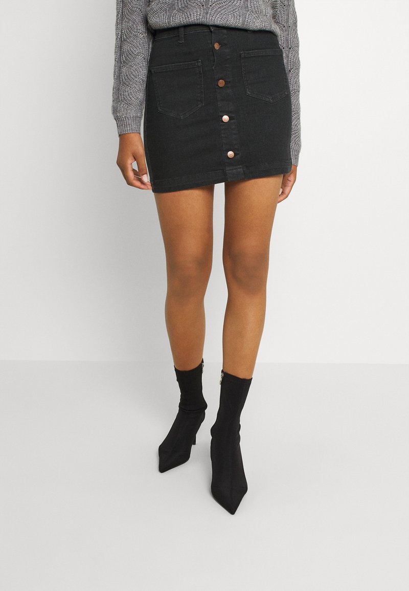 Femme portant une mini-jupe noire boutonnée à l'avant, un pull gris torsadé et des bottines noires à bout pointu avec des fermetures éclair.