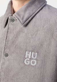 Grå kordjakke med en knappet krave, der har en vertikal ribbet tekstur og broderet "HUGO" logo i lys blå.