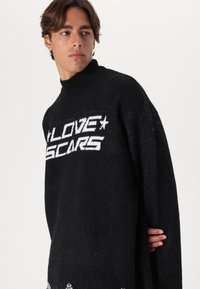 Μαύρο πλεκτό πουλόβερ με ψηλό λαιμό, χαλαρή εφαρμογή και λευκό κείμενο "LOVE SCARS". Διαθέτει υφή στο ύφασμα και ελαφρώς υπερμεγέθη μανίκια.