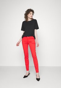 T-shirt preto de manga curta emparelhado com jeans vermelhos justos; modelo usa mules pretos de salto. Tecido liso com detalhes mínimos.