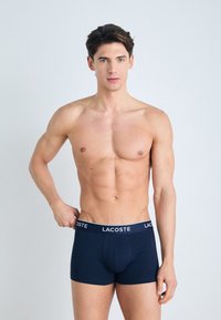 Boxer-briefs bleu marine en tissu doux avec un logo Lacoste blanc sur la ceinture, présentant un design ajusté et une texture lisse.