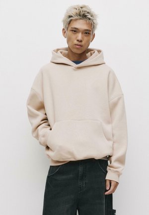 Beige oversized hoodie met een voorzak, capuchon met trekkoord en geribbelde manchetten. Gemaakt in combinatie met zwarte donkere denim broek. Eenvoudige, gladde textuur.