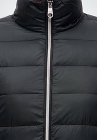 Veste matelassée noire avec une fermeture éclair argentée au centre, montrant un gros plan du haut de la poitrine et de la zone du col.
