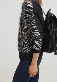 Černý svetr s chlupatými bílými pruhy zebra na rukávech, kombinovaný s tmavými džíny. Součástí je černý batoh s třásněmi.