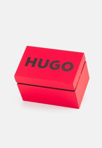Caja de cartón roja con tapa con bisagra, que presenta la palabra "HUGO" impresa en letras negras en negrita en la parte superior. Textura suave.