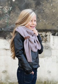 Zwarte leren jas, grijze gebreide sjaal met structuurpatroon, lang blond haar. Donkerblauwe jeans. Voor een verweerde muur.