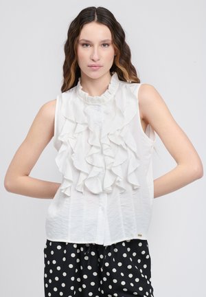 Witte mouwloze blouse met gerimpeld voorpand, ronde halslijn en knoopsluiting. Draagt mee met zwarte broek met witte stippen. Soepele textuur.