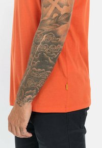 Oranje T-shirt met korte mouwen en een ronde hals, gecombineerd met donkere broek. Opvallende tatoeages zichtbaar op de onderarm; klein geel label aan de zoom.