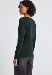 Donna con capelli ricci che indossa una maglietta a maniche lunghe di un verde scuro e jeans neri scoloriti, rivolta di lato con un'espressione neutra.