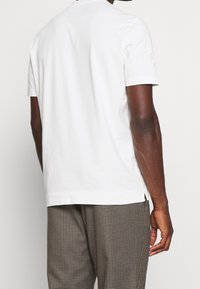Camiseta de algodón blanca con mangas cortas, cuello redondo y aberturas laterales. Combinada con pantalones marrones a cuadros, que exhiben una textura suave y un diseño sencillo.