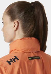 Oranje waterdichte jas met een hoge kraag, voorzien van een zwart logo op de linker schouder en een zwart-groene accenten op de achterkant.