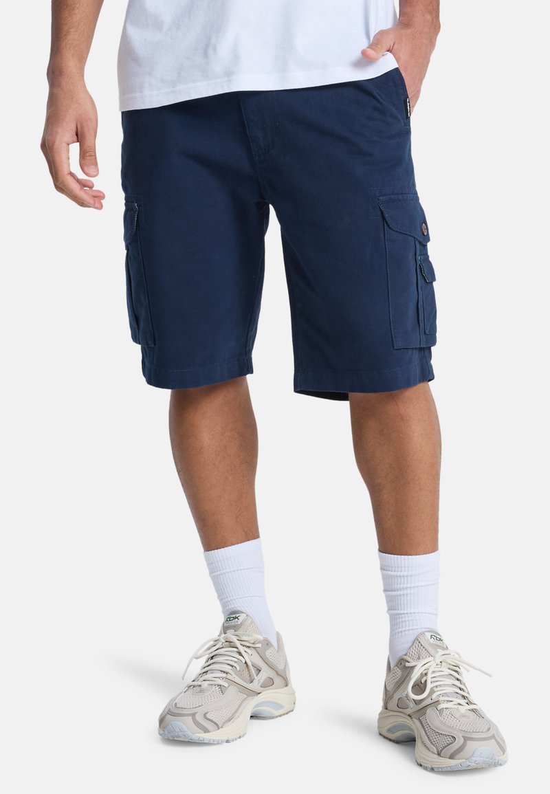 Pantalones cortos cargo de color marino en tejido ligero, con bolsillos laterales y dobladillos enrollados. Combina con zapatillas deportivas grises y calcetines deportivos blancos.