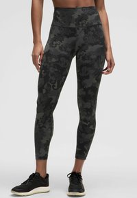 Leggings visoke struka s tamnim vojničkim uzorkom, izrađene od rastezljivog materijala s glatkom teksturom. Upareni s crnim sportskim cipelama.