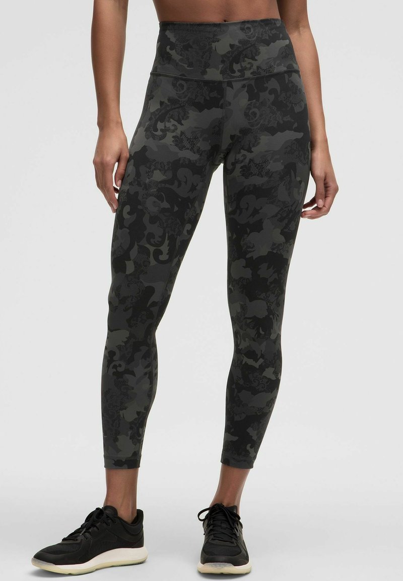 Leggings visoke struka s tamnim vojničkim uzorkom, izrađene od rastezljivog materijala s glatkom teksturom. Upareni s crnim sportskim cipelama.