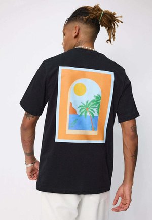 Homme avec des dreadlocks portant un t-shirt noir orné d'un design de fenêtre coloré représentant le soleil, des palmiers, la mer et un paysage montagneux.