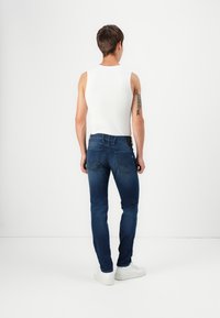 Jeans de mezclilla en azul oscuro con un corte ajustado, combinados con una blusa sin mangas blanca. Con bolsillos traseros visibles y un acabado liso y limpio.