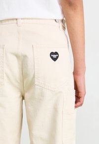 Bež cargo hlače s opuštenim krojem, s dva stražnja džepa i crnim zakrpom u obliku srca s logom "Carhartt WIP".
