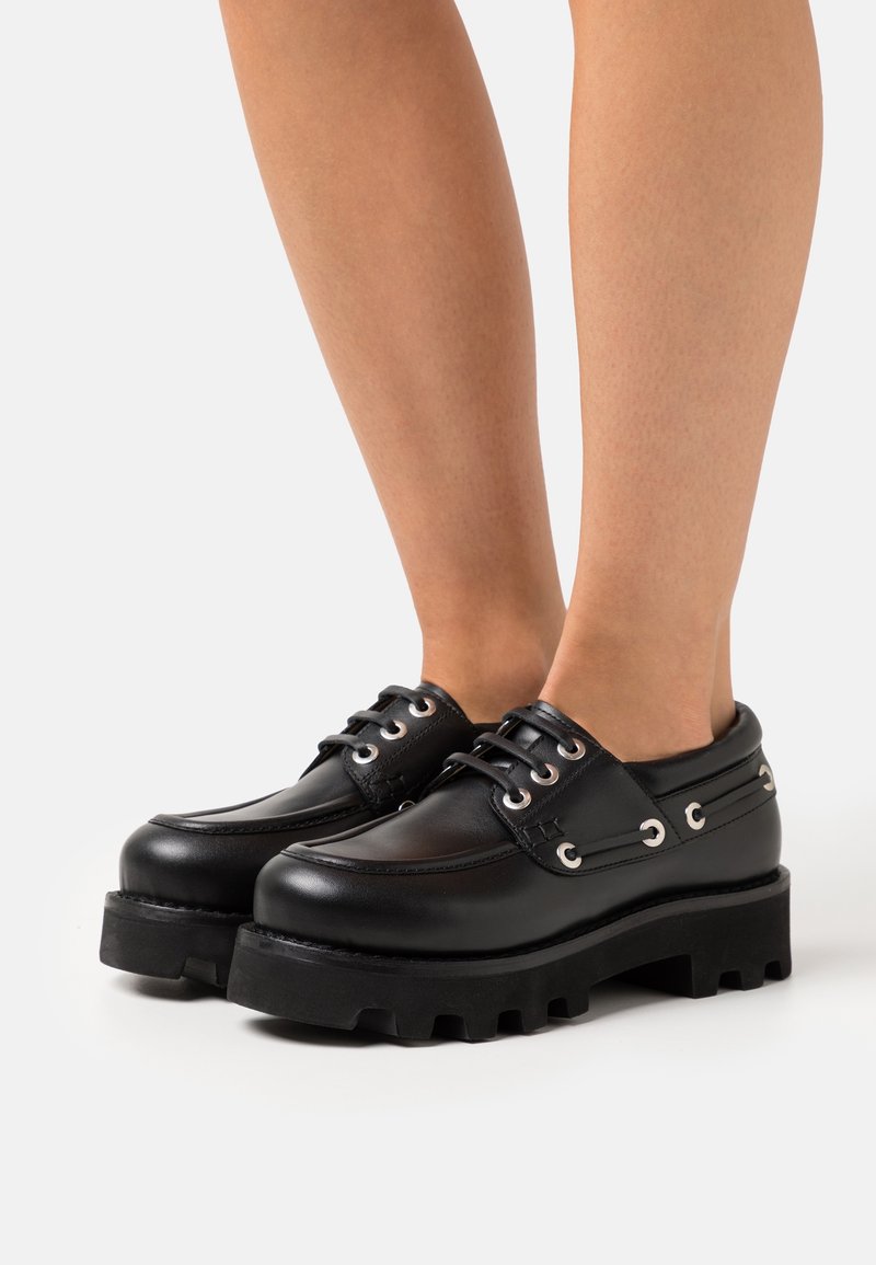 Jambes portant des chaussures en cuir noires épaisses à lacets avec des semelles épaisses crantées, debout sur un fond clair uni.