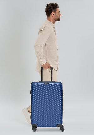 Uomo vestito con un completo beige che cammina con una valigia rigida blu su ruote, tenendo il manico esteso contro uno sfondo chiaro e semplice.
