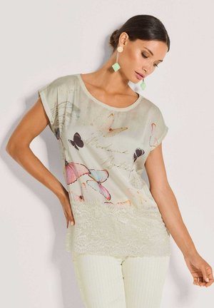 Frau trägt hellbeige Schmetterlings-Print-Bluse mit Spitzenbesatz und weiße strukturierte Hose, posiert mit einer Hand in der Hüfte vor einfarbigem Hintergrund.