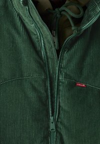 Zielona kurtka ze sztruksu z zamkiem na przodzie, teksturowanymi pionowymi paskami oraz małą czerwoną metką marki przy linii zamka.