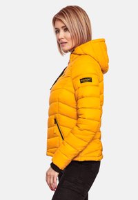 Yellow puffer jacket with a hood, quilted design, black zipper accents, and branded patch on the sleeve. Worn with black pants.