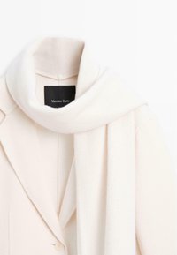 Bílý vlněný kabát s šálovým límcem a odpovídající šálou. Jednoduchý design s jedním zapínáním na knoflík. Měkká textura a minimalistický styl.