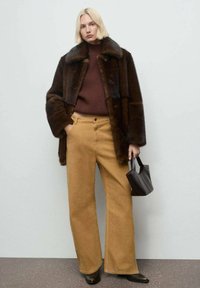 Blondinka v temnosrnem krznenem plašču, burgundski pulover, široke camel hlače, črne škornje, drži majhno temnosrno torbico, stoji ob sivi steni.