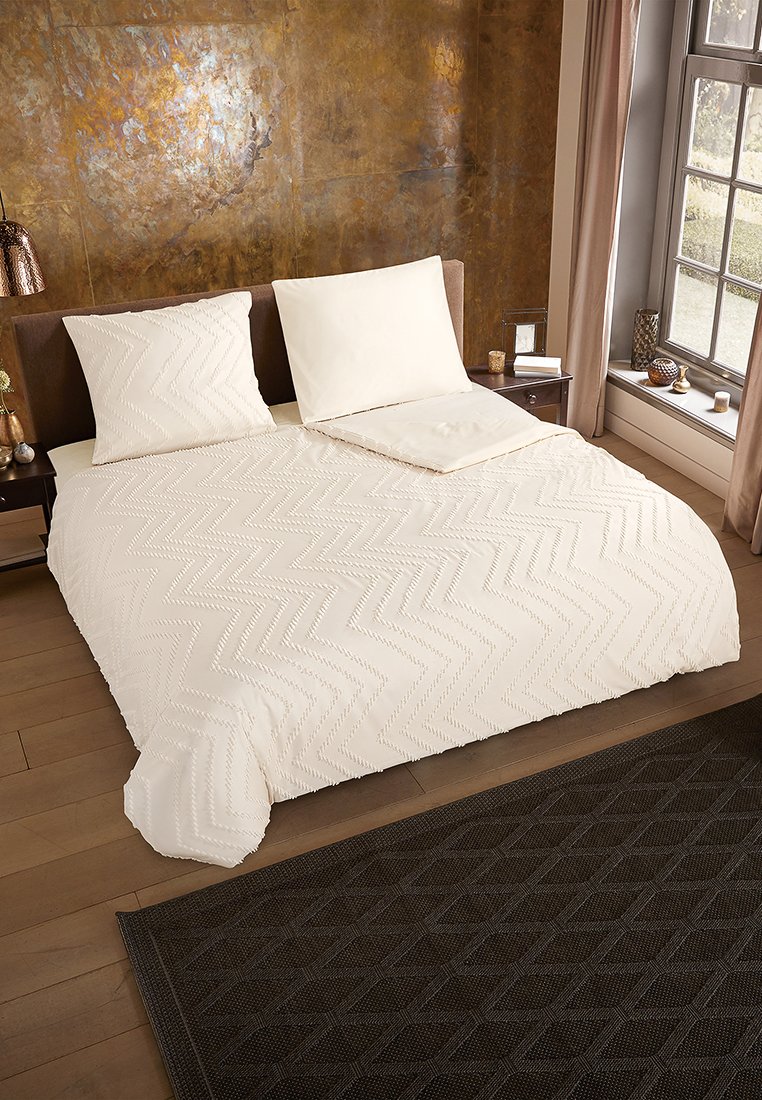 Weißes Bettwäscheset mit einem Zickzackmuster, begleitet von Standardkissen. Dunkel geometrischer Teppich auf Holzboden; bronzefarbene Wand dahinter.