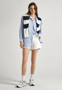 Camicia azzurra chiara con bottoni, pantaloni corti di denim bianchi con orli sfrangiati e un maglione lavorato a maglia a righe blu navy e bianche appoggiato sulle spalle. Calze bianche e sneakers.
