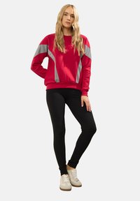 Felpa rossa con dettagli grigi, maniche lunghe e polsini a coste, abbinata a leggins neri e scarpe da ginnastica di colore chiaro.