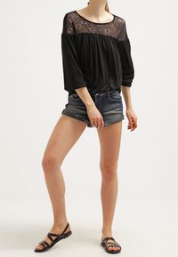 Svart topp med spetsdetaljer vid halsringningen, lös passform, trekvartsärmar, matchad med denimshorts och svarta platta sandaler.