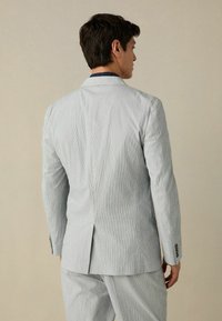 Abito grigio chiaro a righe con tessuto texturizzato, spacco singolo sul retro e due bottoni sulle maniche. Fit sartoriale, design moderno.