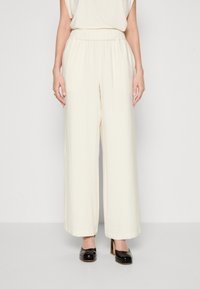 Wijd-uitlopende pantalons in een zachte, gestructureerde crèmekleurige stof. Voorzien van een elastische tailleband en een vloeiende silhouette. Gekleed met zwarte hakken.