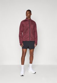 Mørkerød zip-up jakke med geometriske mønstre, lange ærmer og sorte accenter; bæres med sorte shorts og hvide træningssko.