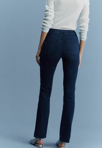 Jeans evazați de culoare albastru închis sunt prezentați din spate. Au două buzunare la spate, un talie înaltă și o textură netedă. Este purtat un pulover light.