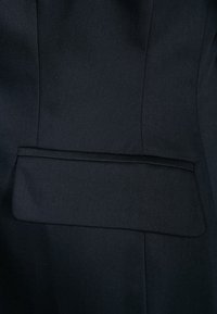 Close-up van donker marineblauw colbertstof, met een horizontale klepzak en zichtbare verticale naadstiksels.