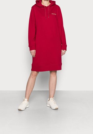 Kvinde iført en rød langærmet hættetrøjekjole med snore og hvide sneakers, stående mod en ensfarvet lys baggrund.