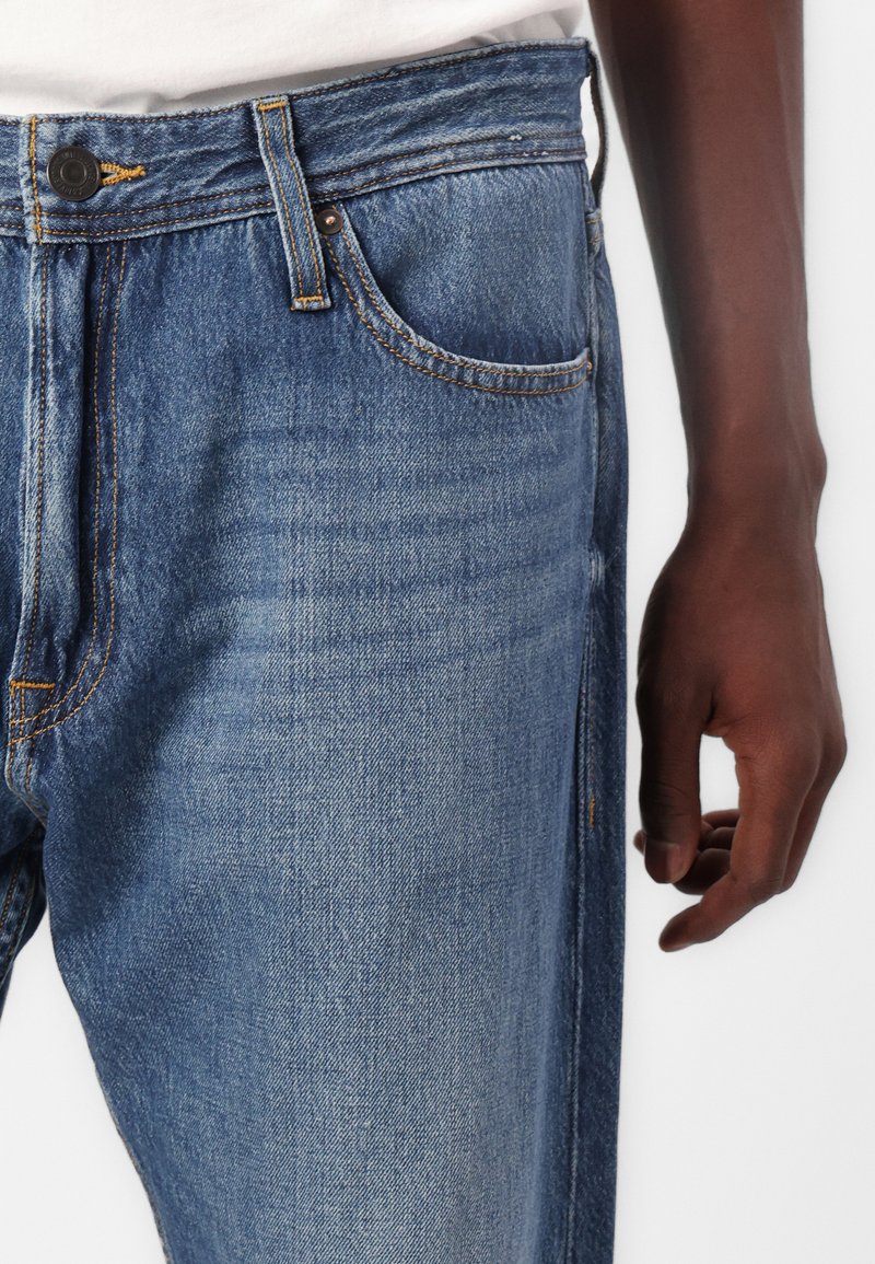 Hellblaue Jeans mit hoher Taille, Five-Pocket-Design, gelben Nähten und Knopfverschluss. Eine Hand liegt an der Seite.