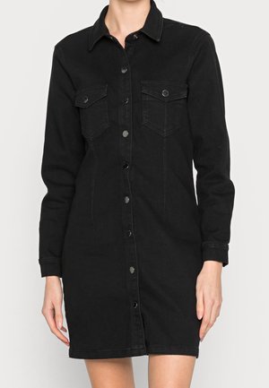Mujer con un vestido vaquero negro de manga larga con botones, bolsillos en el pecho y cuello, de pie frente a un fondo claro y liso.