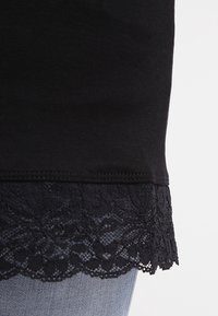 Svart t-shirt med spetsdetalj vid fållen. Mjuk bomullsmaterial med intrikata blommönster i spets. Närbild visar detaljer.