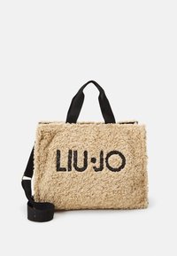 Beżowa torebka typu tote z frędzlami z czarnym logo "LIU∶JO", z krótkimi uchwytami i odczepianym czarnym paskiem. Miękka tekstura i prostokątny kształt.
