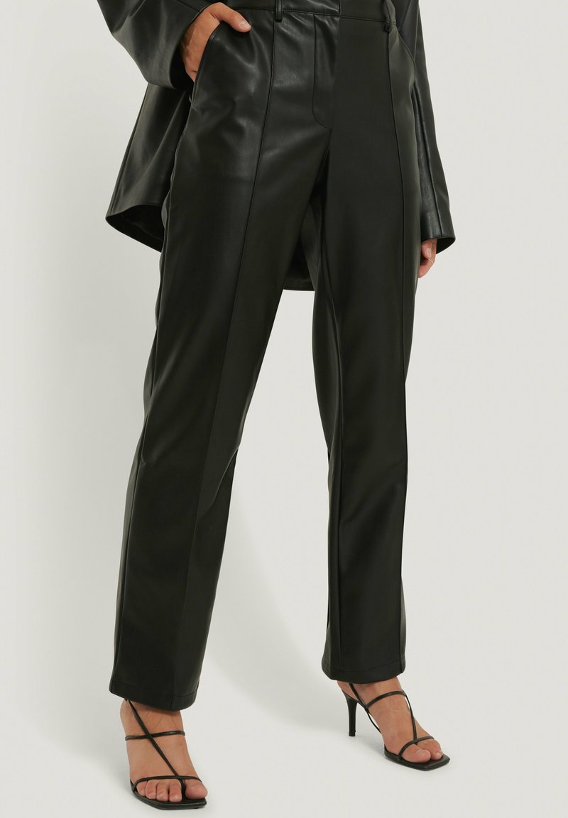 Femme portant un pantalon taille haute en similicuir noir avec des coutures avant et des sandales à talons hauts noires à brides, la main dans la poche.