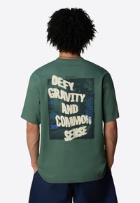 Camiseta de manga corta verde con un audaz gráfico en la parte posterior que dice "DESAFÍA LA GRAVEDAD Y EL SENTIDO COMÚN", rodeado de varias formas geométricas.