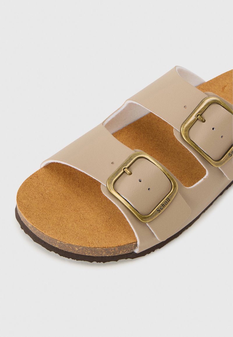 Sandale beige à enfiler avec deux sangles réglables et boucles dorées. Semelle en liège et semelle en caoutchouc texturé. Design minimaliste.